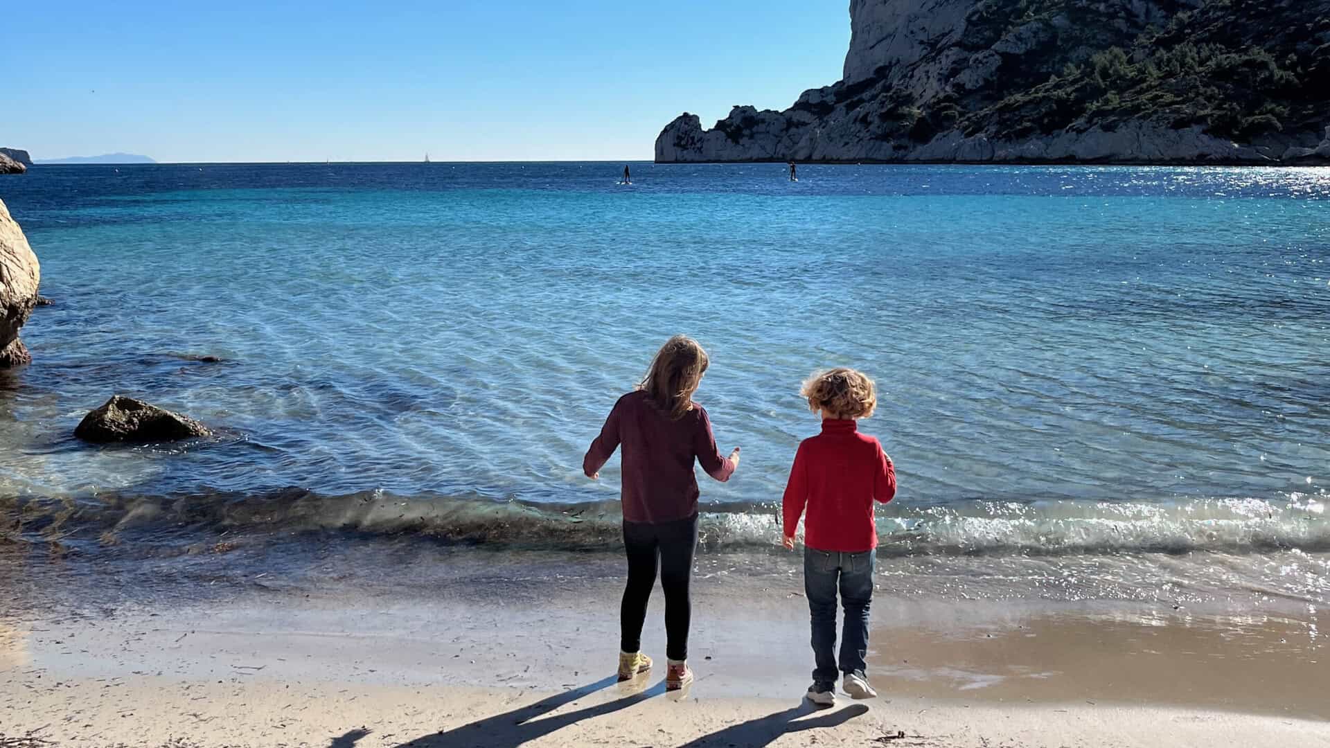 Deux enfants jouent à la plage de Marseille, eaux claires et falaises.